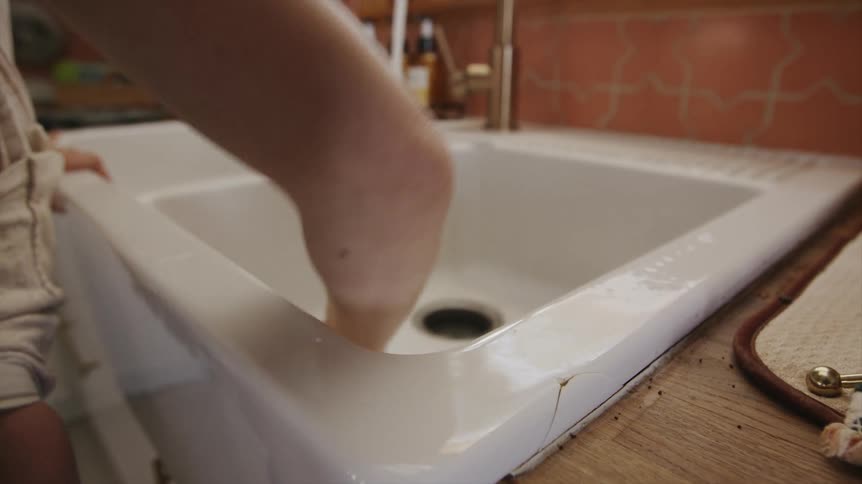 Woman using a cleaning cloth and the tap water to clean a white sink
