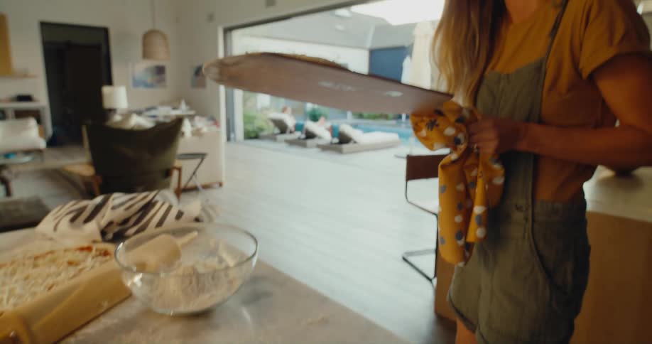 A woman carries a heart shaped pizza from her kitchen to her pool side patio