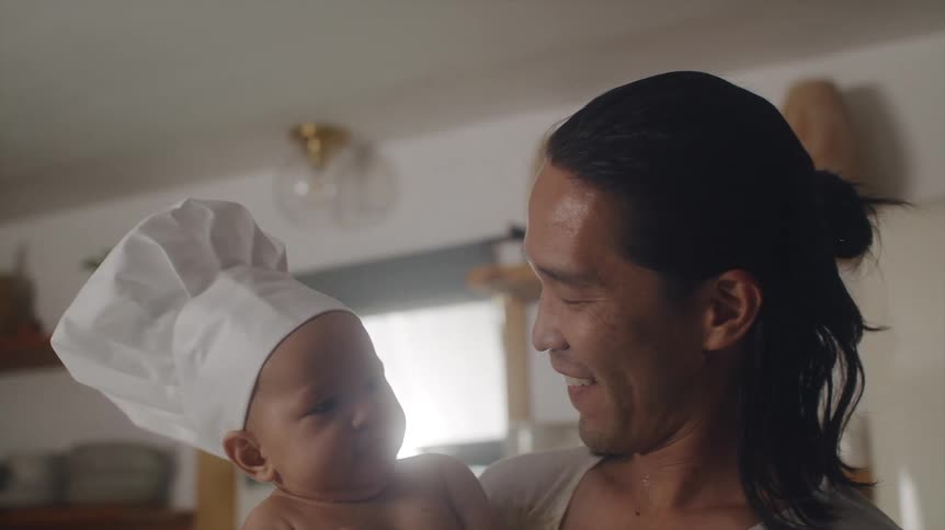 Father with long hair white shirt and apron cleans babys face with towel wearing chef hat in the kitchen