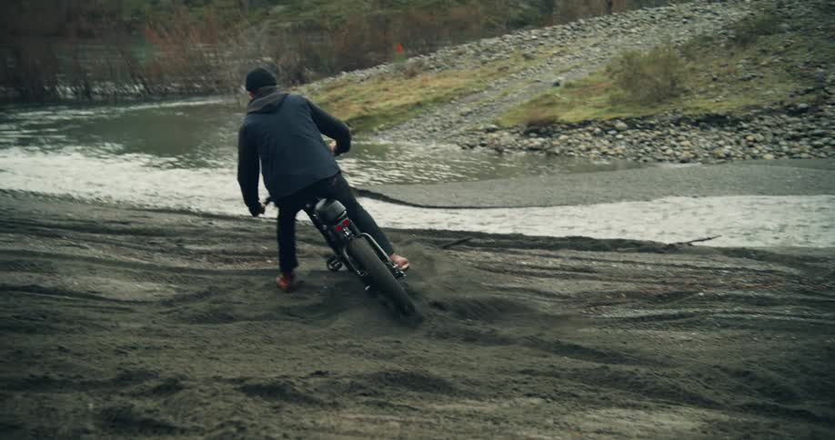 Man with black wooly hat naval blue vest black jacket jeans and pair of brown leather shoes rides dirt bike on river bank