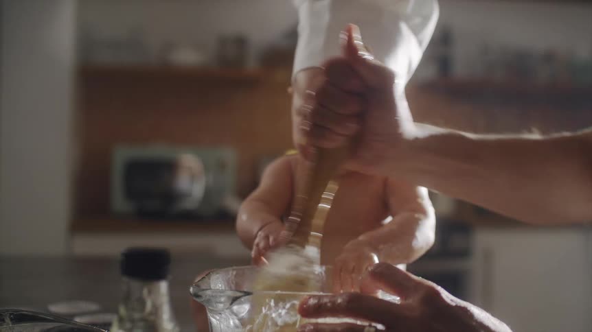 A man prepares food in a mixing bowl as a baby in a chef hat watches from the side