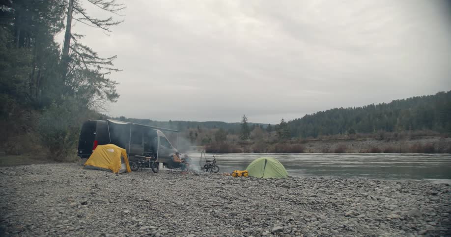 A van is parked next to a lake with tents and people are sitting near a fire