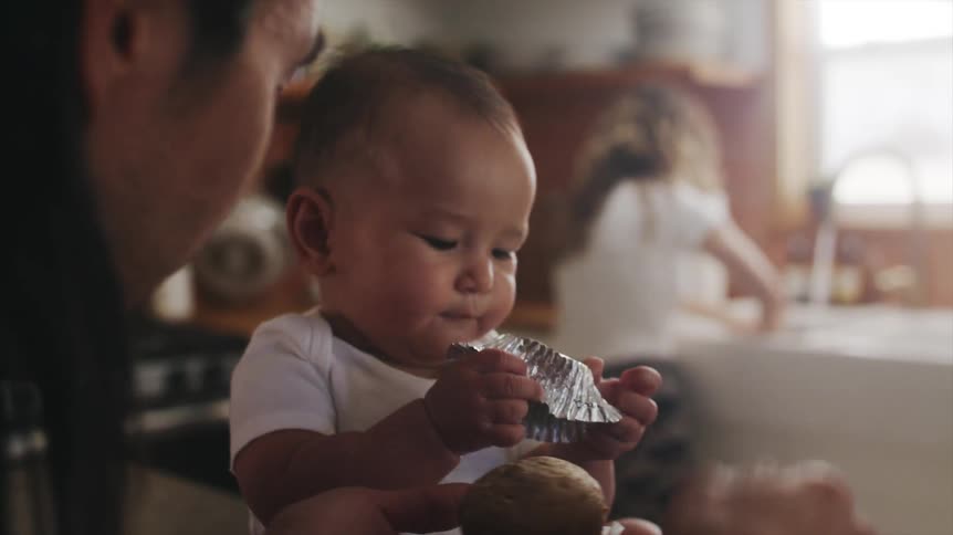 Baby sitting at the kitchen counter eating a candy his father just gave him while the mother is washing the dishes at the sink behind them