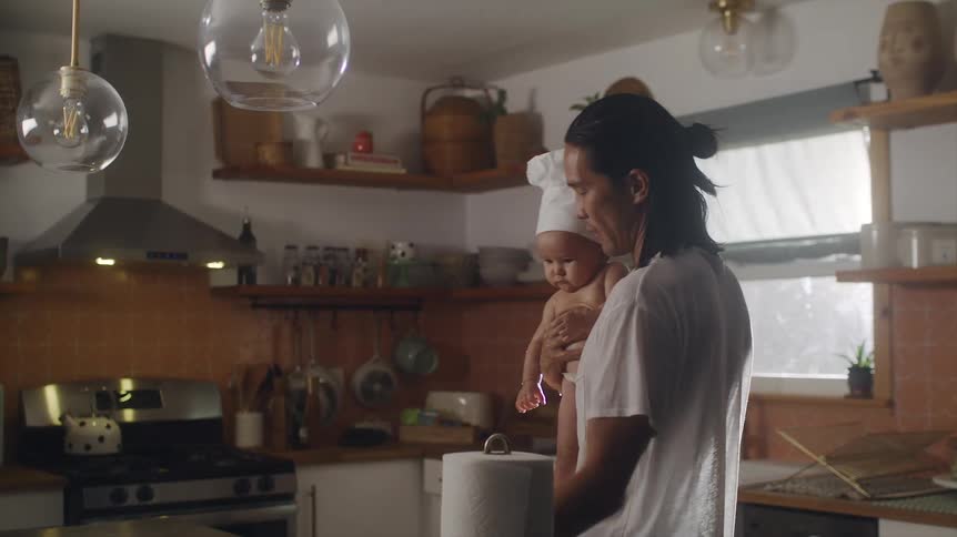 Father with long hair white apron and t shirt holds baby wearing chef hat and wipes kitchen island in the kitchen