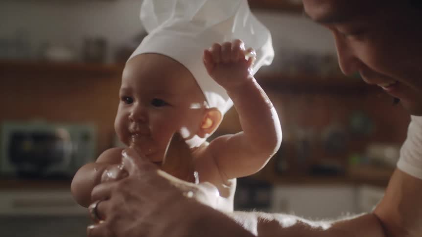 A man helps a baby in a chef hat to mix food in a kitchen during the day