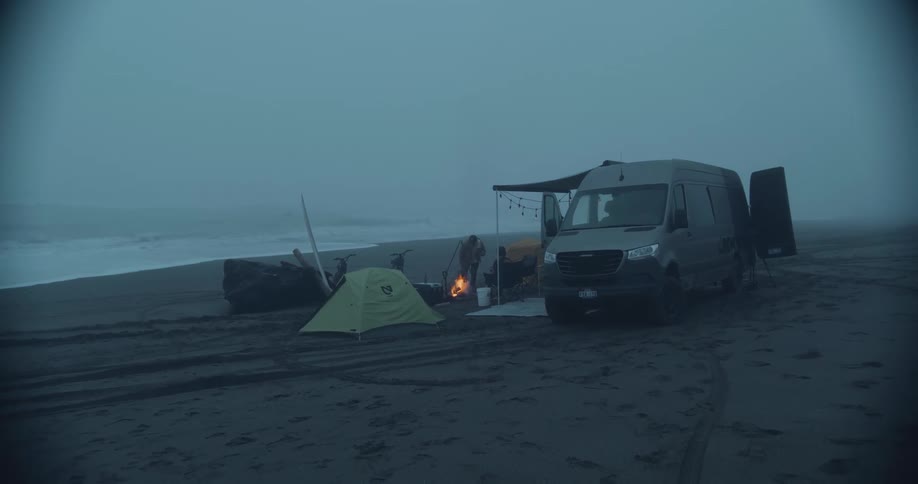 A van is parked along the coast with tents and people walking near a campfire