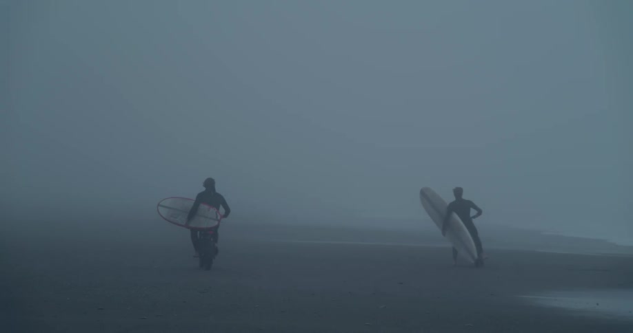 Two surfers carry boards in one arm while steering motorcycles with the other as they ride across beach on densely foggy day