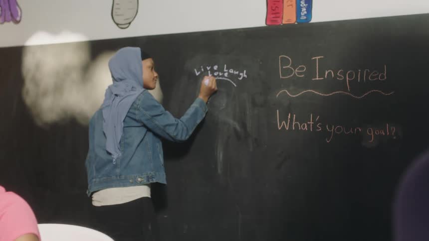 Teenaged girl draws a circle on a chalkboard while another girl sits at a table reading a thick paperback book