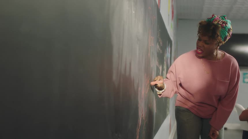A woman speaks while writing on a blackboard in a classroom
