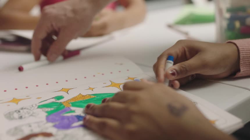 Children color in coloring books while a teacher stands above them