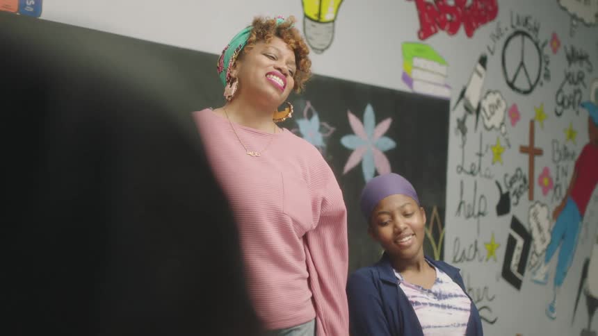 Teacher with green headband earrings and pink sweater talks to students wearing headscarves and headbands in classroom