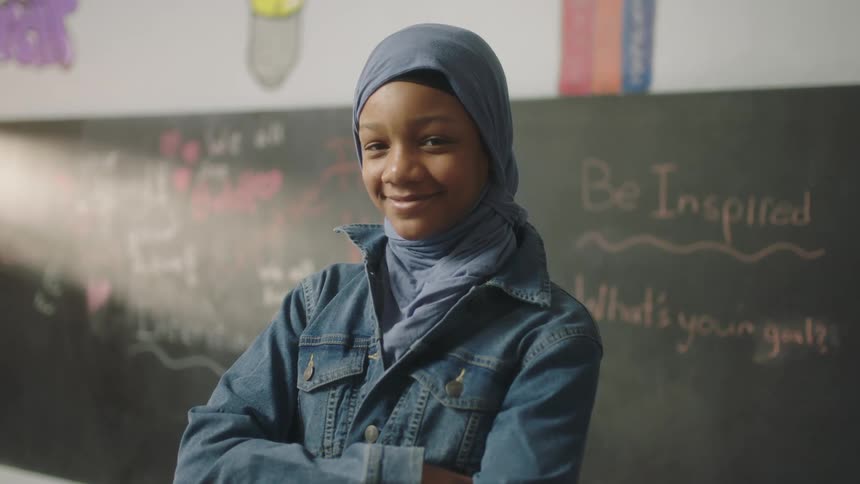 A young woman smiles into the camera while standing before a blackboard in a classroom