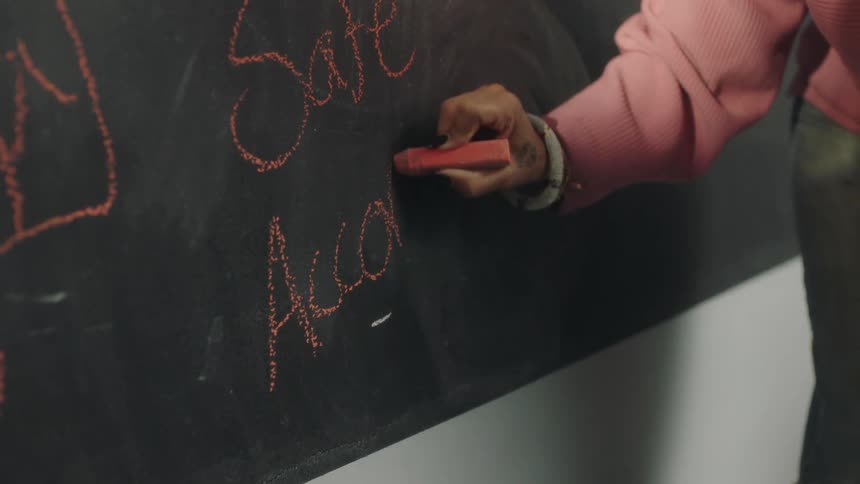Woman in a pink sweater writes on a chalkboard while two women sit in chairs at a table
