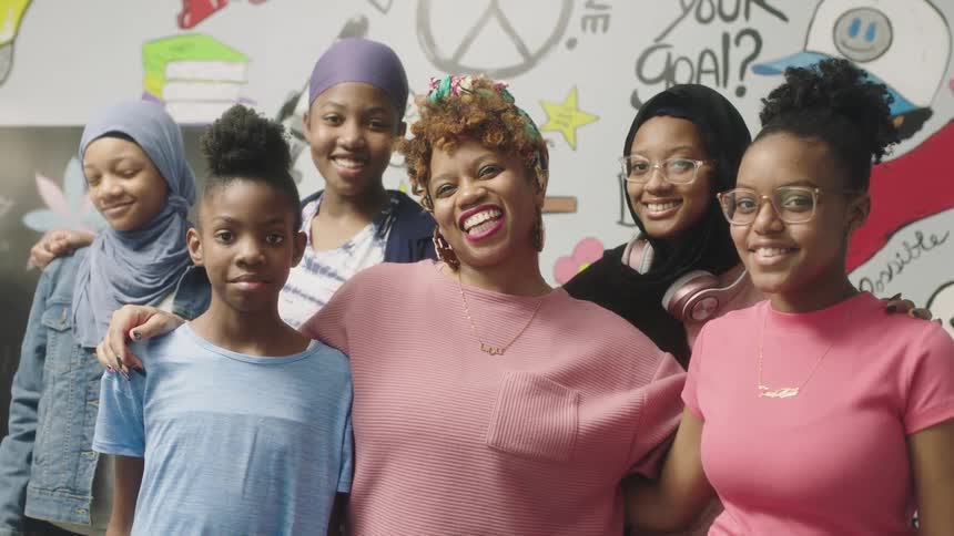 Female teacher with headband earrings and pink shirt stands among students posing for camera in classroom at school