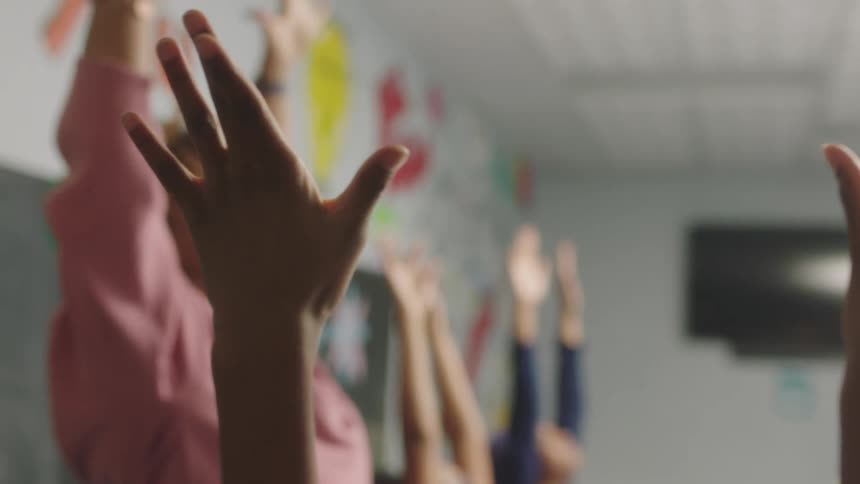 Teacher wearing green headband earrings and pink sweater raises hands with students in the classroom