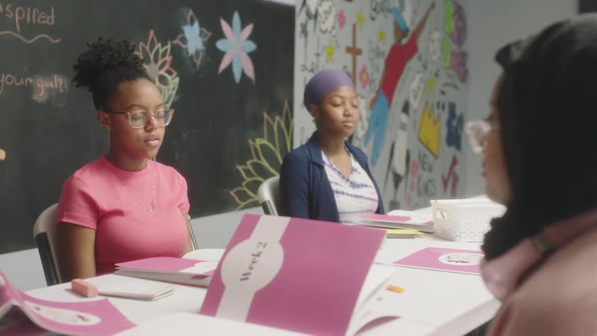 Three teenaged girls breathe in deeply then exhale as they sit around a table that is covered with pink workbooks