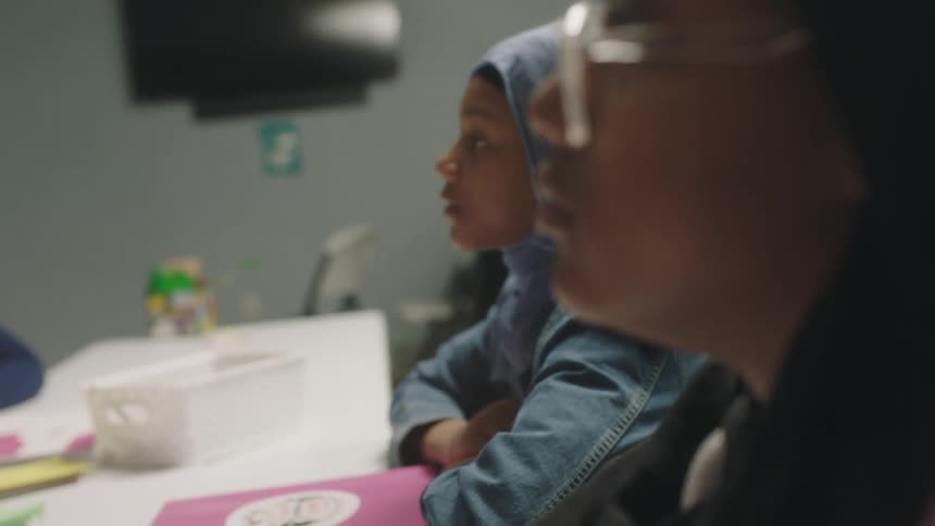 Female students with glasses denim jacket and headscarves sit at desk and listen to teacher in classroom