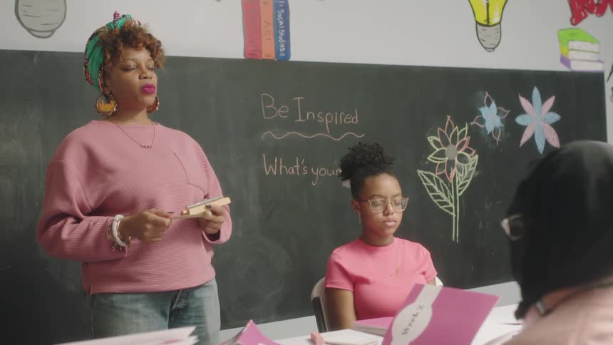Students sit in desks with their eyes closed and listen to their teacher who stands at the front of the class and talks with her eyes closed