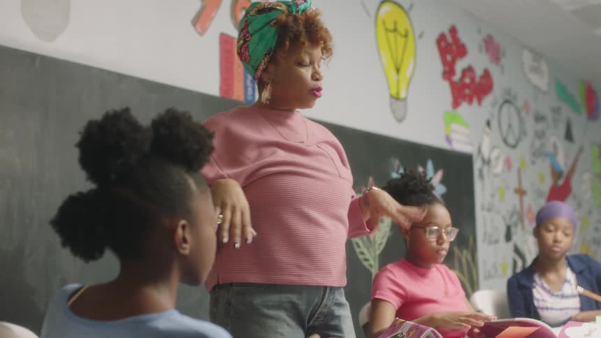 Woman in a pink sweater talks while standing in front of a chalkboard while three women sit at a table in front of her