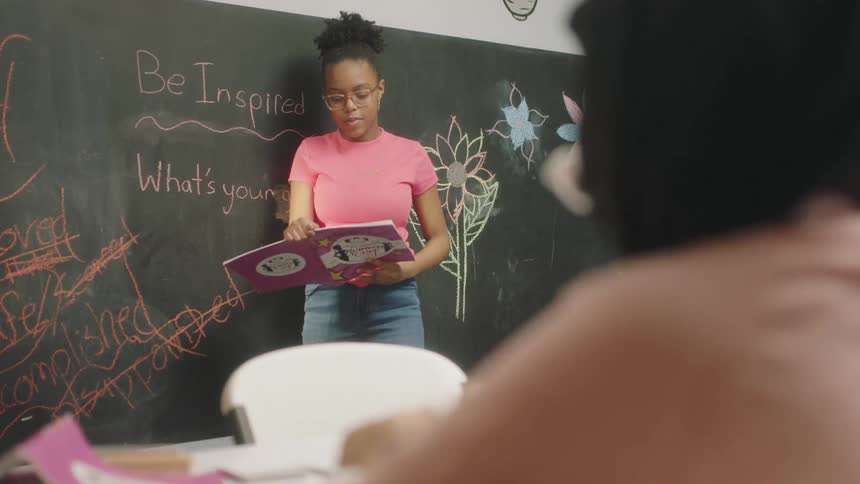 Student stands near blackboard reading from a book