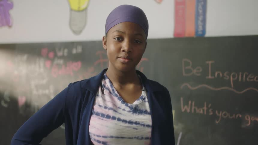 A teenager stands in front of a chalk board at school and smiles