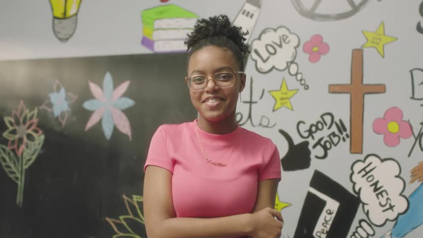 Female student with curly hair glasses earrings and pink t shirt faces the camera with eye contact smiling in classroom at school
