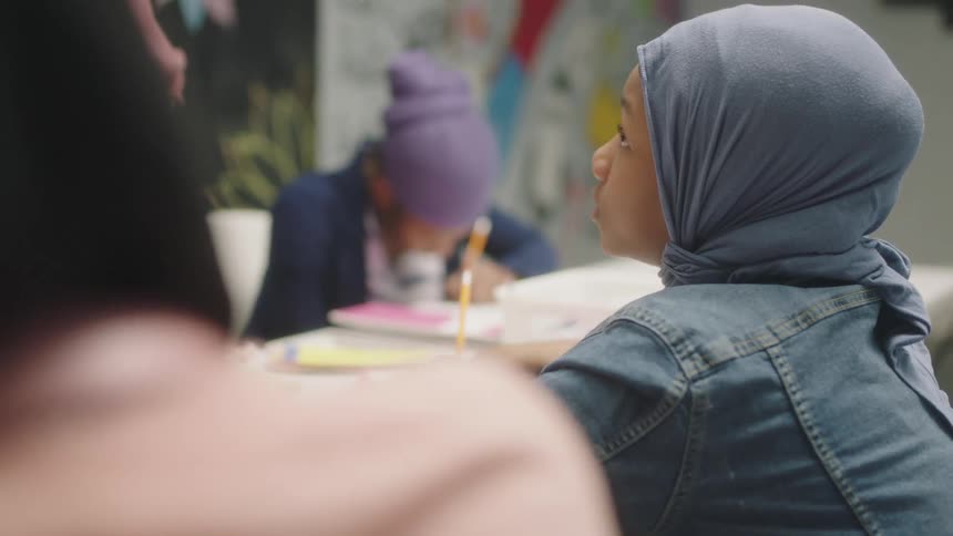 Students sit at desks in a classroom and write and talk as the teacher stands in front of them