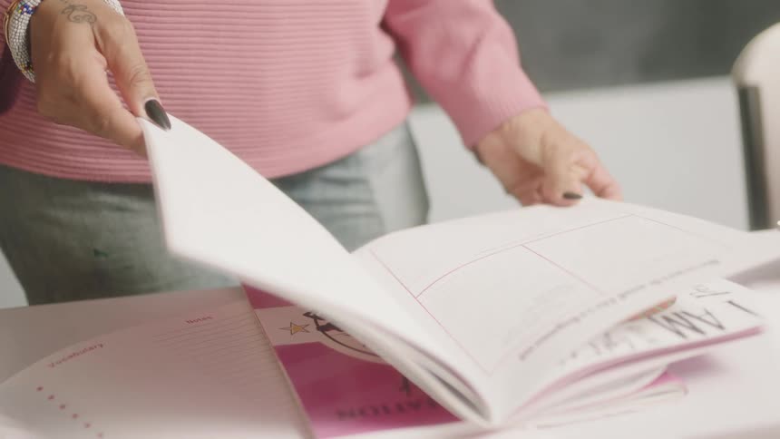 Woman in a pink sweater turns the pages of a book while standing in front of a black chalkboard