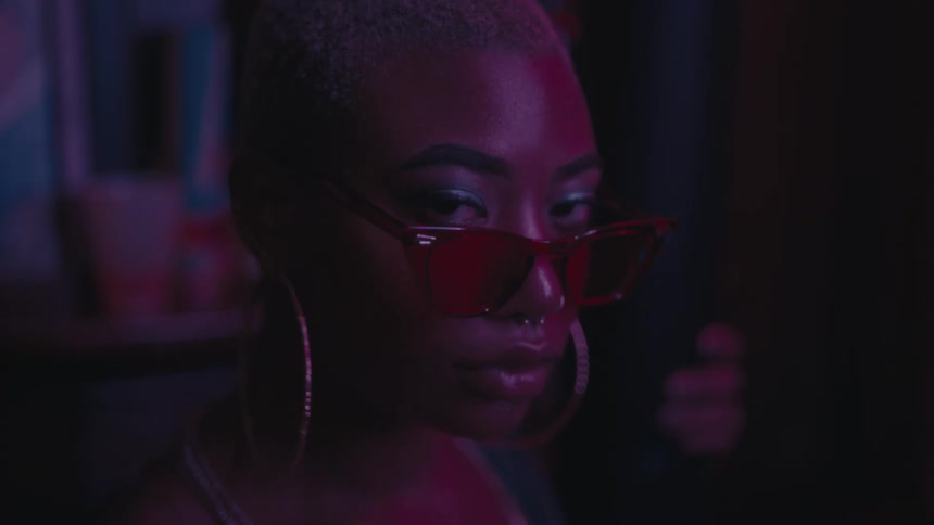 A black woman with large hoop earrings wears sunglasses in a dark room