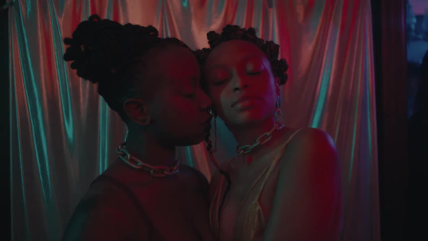 Two women cuddle together posing in front of metallic curtain with people in background in club