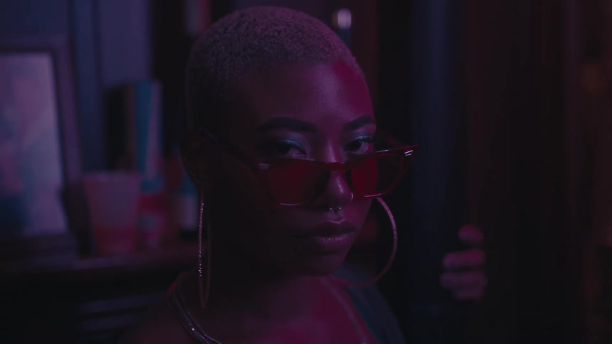 A black woman with very short hair poses for the camera while wearing sunglasses in a darkened room