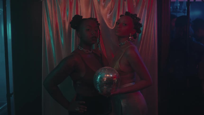 Black woman holds a shiny sphere while standing in front of a shiny curtain with another black woman