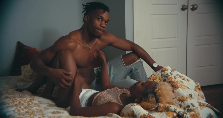 Shirtless man with dreadlocks and gray pants sits on bed touching womans leg lying on bed in bedroom