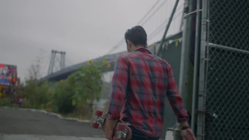 A man carrying a skateboard walks in an urban area near a bridge