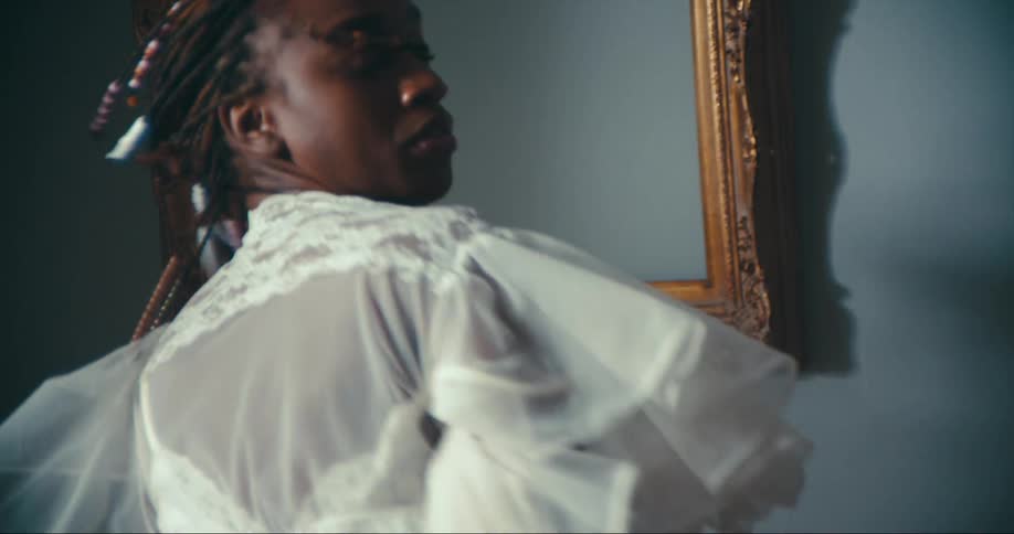 Woman with dreadlocks and white dress dances in front of wall with picture frame