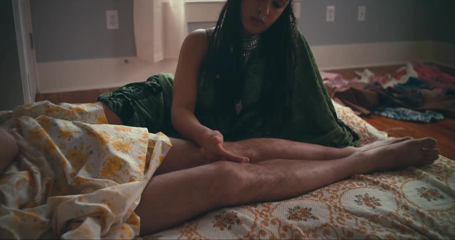 Woman with green dress dreadlocks sits on bed and strokes mans legs lying on bed in bedroom
