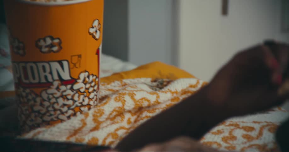 A woman eats popcorn from a large cup while lying in bed watching her tablet