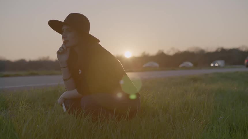 Woman kneeling in the grass next to a road at sunset
