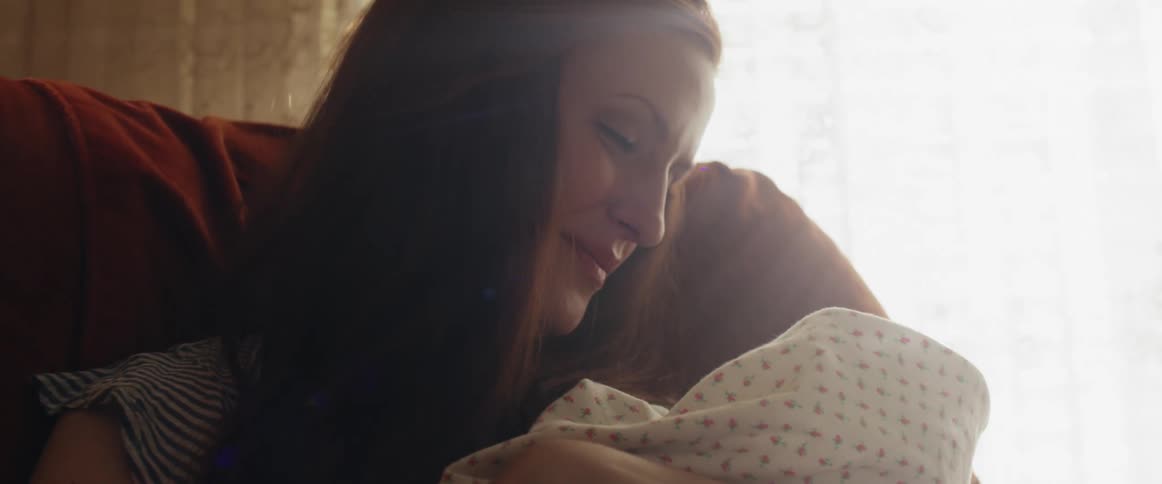 A woman cradles a baby wrapped in a blanket while sitting indoors during the day