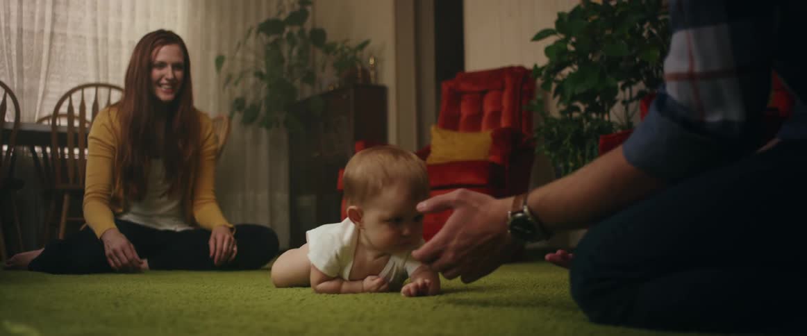 Mom smiles joyfully as baby crawls across the carpet to dad who is waiting with open hands
