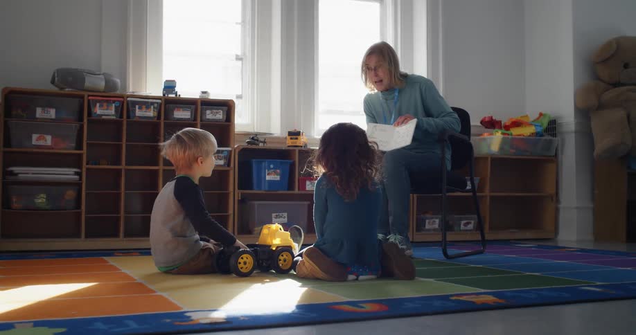 Two little children sit on a mat listening to a teacher read a book