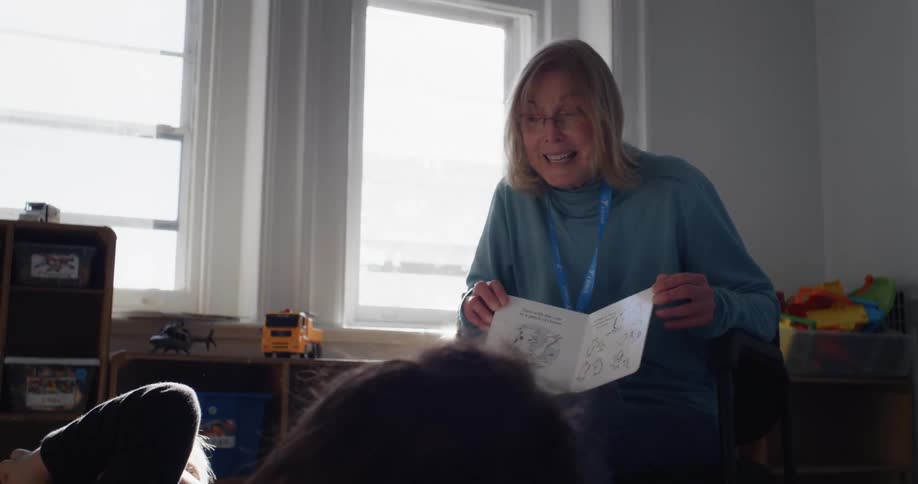 A woman reads a picture book to a pair of children sitting on the floor in front of her
