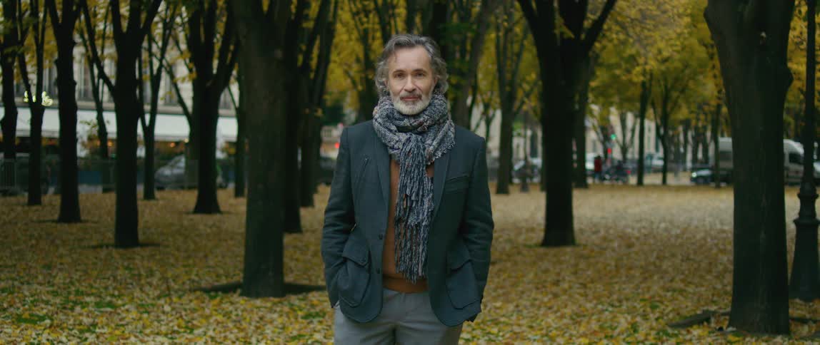 A man wearing a coat and a scarf around his neck is standing in a park with trees behind him and yellow leaves on the ground