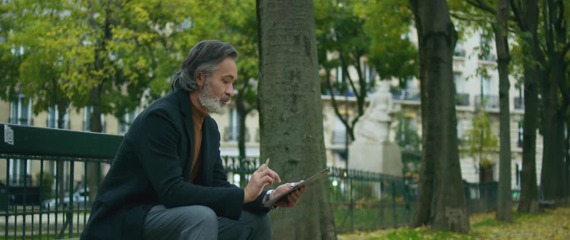 Man sits on bench in park scrolling on tablet as cars pass on street in the background