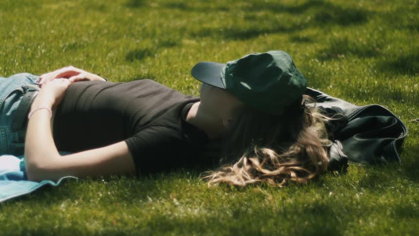 A woman lying in the grass with a cap over her face napping in the sun