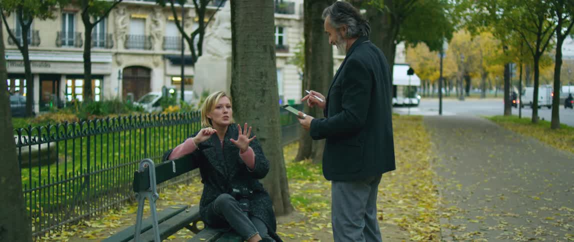 A man is standing near a blond woman sitting on a park bench and she is moving her hands as she talks to him