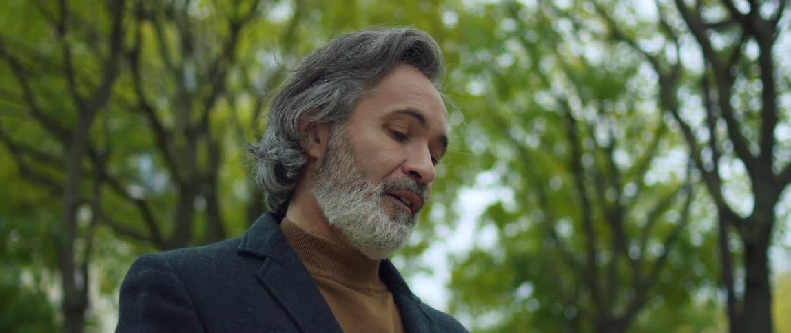 A man is standing among trees looking down at something and smiles