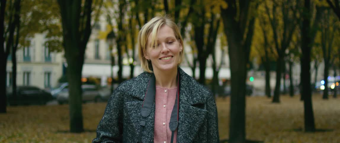 A woman with blond hair is standing in a park with trees behind her and smiling