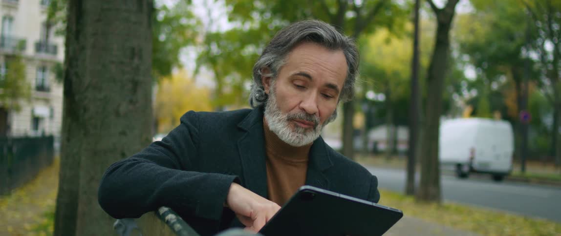 A man is sitting on a park bench holding an electronic tablet and a pen as he looks around