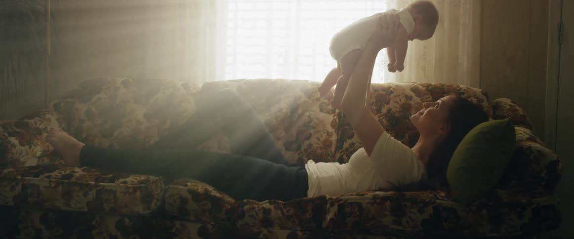 Mother lies on her back on the couch playing by holding baby over her as bright sun shines through the window